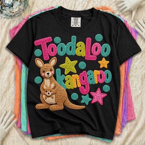 Può includere: T-shirt nero con una grafica colorata raffigurante un canguro e le parole "Toodaloo Kangaroo". Il design include stelle e cerchi rosa, gialli e turchese. Il canguro è marrone con un bambino nella sua tasca.