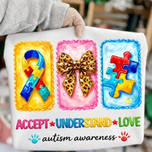 Op de afbeelding: Wit T-shirt met drie ingelijste afbeeldingen: een lint, een strik en puzzelstukjes. De tekst luidt "ACCEPT * UNDERSTAND * LOVE" en "autism awareness" met handafdrukken. Het lint is geel, blauw, rood en groen.