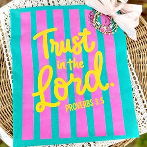 May include: A teal and pink striped fabric with the words "Trust in the Lord" in yellow script. The text "Proverbs 3:5" is below. A collection of bracelets and a pink bow are in the upper right corner.