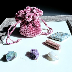 May include: A small, round, pink velvet drawstring pouch with a textured, crocheted surface. Beside the pouch are four distinct stones: a flat blue square, a cylindrical pink stone, a rough purple rock, and a clear, pointed crystal.