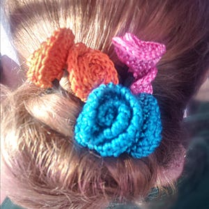 May include: A close-up of a hairstyle featuring several handmade crochet hair accessories. The accessories are in vibrant colors: orange, pink, and turquoise. The hair is a light brown color.