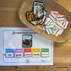 Pode incluir: Jogo educativo com cartões e um quadro. O quadro diz "Je construis ma phrase" e tem espaços para palavras. Os cartões incluem imagens de uma girafa e um caminhão. Uma bandeja de madeira contém cartões de palavras adicionais.