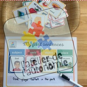 May include: Educational game with sentence-building cards and a whiteboard. The cards feature images and words like "apple" and "magazine." The whiteboard has the text "My first sentences" and example sentences. A black marker is also present.