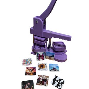 May include: A purple button maker with a lever, and several square buttons with various designs. The buttons feature images of a butterfly, a sunflower, people, and text that reads "Be the reason someone smiles today!" and "Sunshine".