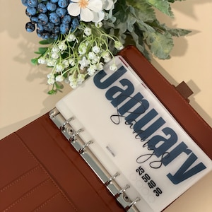 May include: A brown leather bound planner with a clear plastic insert that says "January" in blue lettering. The insert also has a grid of numbers for the month.