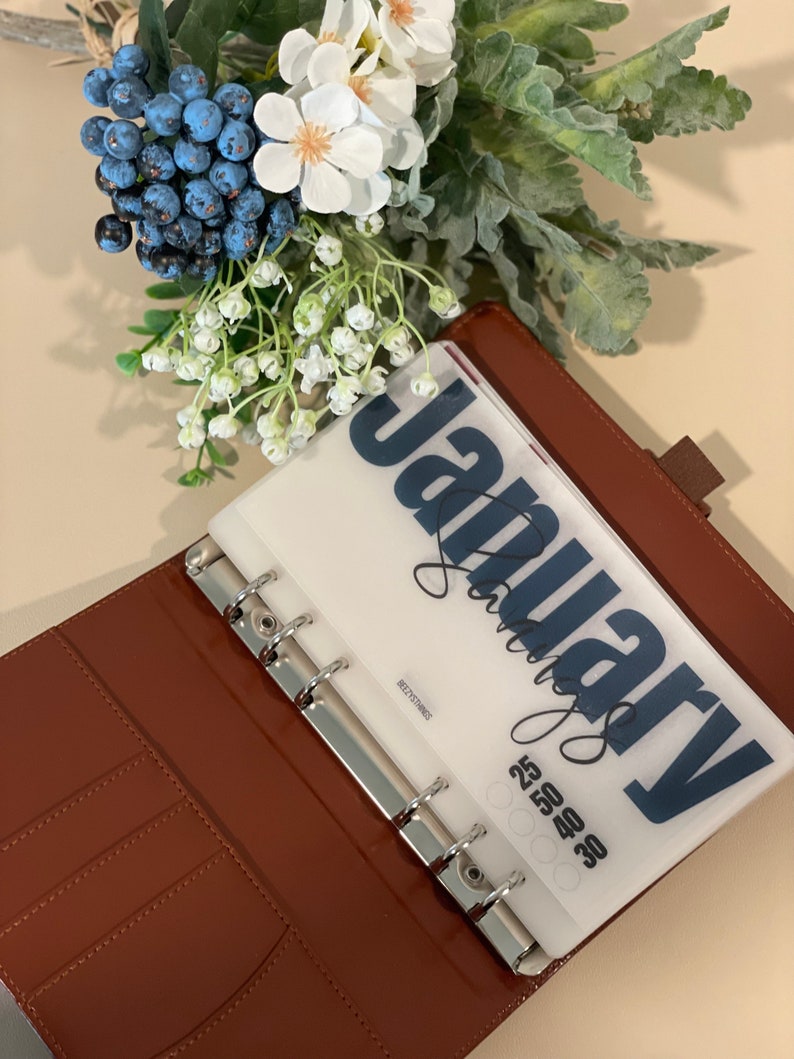 May include: A brown leather bound planner with a clear plastic insert that says "January" in blue lettering. The insert also has a grid of numbers for the month.