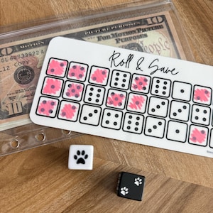 May include: A white card with the text "Roll & Save" and a grid of dice faces, some marked with pink. Two dice, one white with a paw print and one black with paw prints, are on a wooden surface. A clear plastic bag with money is in the background.