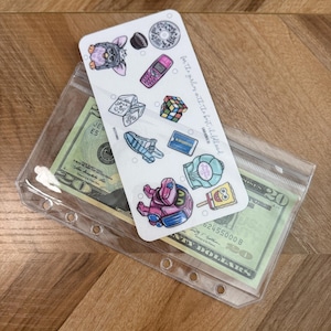 May include: A clear plastic pouch containing a twenty-dollar bill and a sticker sheet. The sticker sheet features various nostalgic images, including a Furby, a Rubik's Cube, and a pink flip phone. Text on the sticker sheet reads "for the girlies with the best childhood."