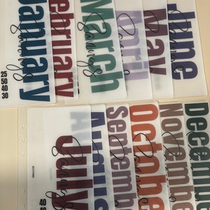 May include: Twelve clear plastic sheets with the months of the year printed on them in bold, colorful lettering. Each sheet has a different color and the month name is written in cursive at the bottom.