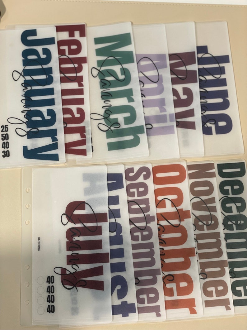 May include: Twelve clear plastic sheets with the months of the year printed on them in bold, colorful lettering. Each sheet has a different color and the month name is written in cursive at the bottom.