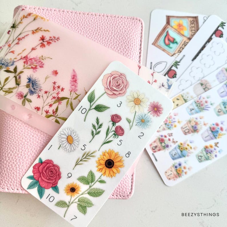 Puede incluir: Una cubierta de agenda rosa texturizada con una superposici&oacute;n floral transl&uacute;cida, acompa&ntilde;ada de varias hojas de pegatinas. Las pegatinas muestran diversas ilustraciones bot&aacute;nicas, incluyendo rosas, margaritas, girasoles y arreglos florales en macetas.