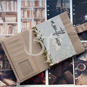 May include: A beige leather bound planner with a clear plastic insert that says "month ahead" in black script. The planner is open and lying on a table with books and other items.