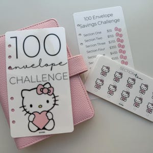 Può includere: Un'agenda rosa con una carta che dice "100 envelope CHALLENGE" e presenta una grafica di Hello Kitty che tiene un cuore rosa. Le carte aggiuntive includono un elenco di sfide di risparmio e una carta con grafiche di Hello Kitty.