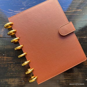 May include: A brown planner with gold-colored disc binding and a matching tab closure. The planner has a textured cover and is closed. The planner is on a dark wood surface.