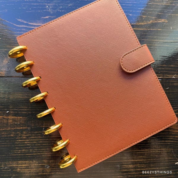 May include: A brown planner with gold-colored disc binding and a matching tab closure. The planner has a textured cover and is closed. The planner is on a dark wood surface.