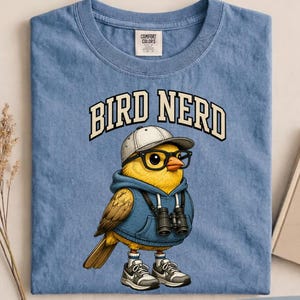 Op de afbeelding: Een blauw t-shirt met de tekst "BIRD NERD" boven een cartoonvogel met een bril, een hoodie, een pet en sneakers. De vogel heeft een verrekijker. Het shirt is gelabeld "BLUE JEAN".