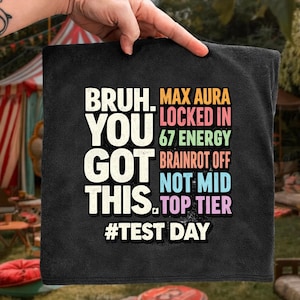 May include: Black square towel with the text "BRUH. YOU GOT THIS. #TEST DAY" in white, and "MAX AURA LOCKED IN 67 ENERGY BRAINROT OFF NOT MID TOP TIER" in various colors. The towel is held up by a hand.