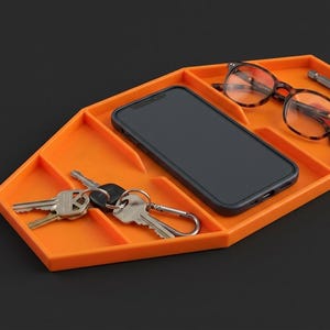 May include: An orange, multi-compartment tray with a hexagonal shape. The tray holds a black smartphone, a pair of tortoiseshell-patterned eyeglasses, a metal screwdriver bit, and a set of silver-colored keys attached to a metal carabiner.