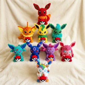 Puede incluir: Una colección de figuras de Pokémon hechas a mano con ganchillo en varios colores. Las figuras incluyen personajes rojos, naranjas, amarillos, verdes, azules, morados y rosas, cada uno sosteniendo una Poké Ball roja y blanca. Las figuras están dispuestas sobre una superficie blanca y suave.