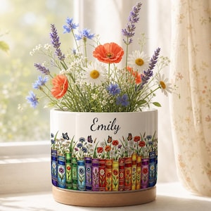 May include: A white ceramic planter with a wooden base, featuring a colorful illustration of books and wildflowers around the exterior. The name Emily is printed in black script on the front. The planter is filled with lavender, poppies, and daisies.
