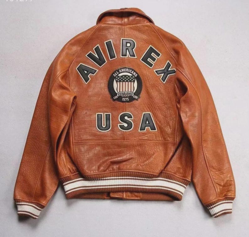 May include: Brown leather jacket with the text "AVIREX" and "USA" on the back. The jacket has a round patch with the words "ALL AMERICANS" and "ESTABLISHED 1975". The jacket has white and brown striped cuffs.