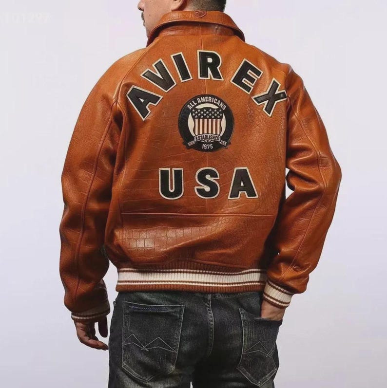 May include: Brown leather jacket with the word "AVIREX" across the shoulders and "USA" at the bottom. The jacket has a patch with the words "ALL AMERICANS ESTABLISHED 1975". The jacket has a ribbed collar and cuffs with white and brown stripes.