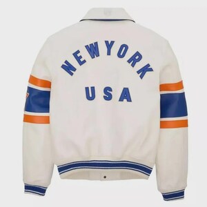May include: Cream-colored jacket with the word "NEW YORK" arched across the back in blue lettering, with "USA" underneath. The sleeves have blue and orange stripes. The jacket has a collar and a blue and white striped trim.