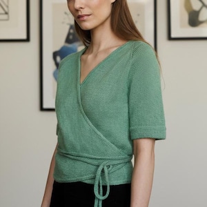 May include: A woman wearing a light green wrap top with short sleeves and a tie at the waist. She is standing in front of a wall with framed abstract art. The top is paired with black bottoms.