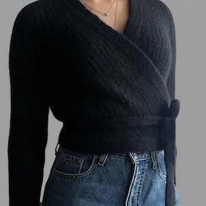 May include: A black wrap sweater with long sleeves and a tie closure at the waist. The sweater is made of a textured knit fabric and is paired with blue denim jeans.