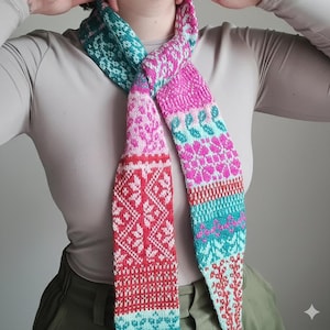 May include: A colorful knitted scarf with a geometric and floral pattern. The scarf features a combination of pink, red, teal, and white. The scarf is made of soft yarn and is designed to be worn around the neck. The scarf is a handmade accessory.