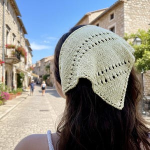 May include: A light green knitted headscarf with a geometric pattern. The headscarf is worn on the head, with the back of the head and hair visible. The background shows a cobblestone street and buildings.