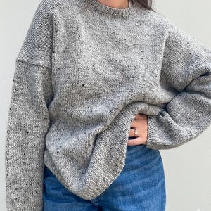 Może przedstawiać: Szary, nakrapiany sweter z dzianiny z okrągłym dekoltem. Sweter ma luźny krój i długie rękawy. Tkanina ma czarne i brązowe plamki. Osoba ma na sobie niebieskie dżinsy.