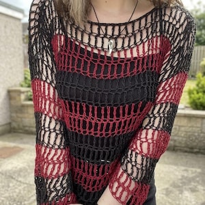 Peut inclure: Un pull à mailles ouvertes rayé noir et rouge. Le pull a une encolure ronde et des manches longues. Un pendentif argenté est suspendu à un cordon noir. Le pull est porté sur un haut noir.