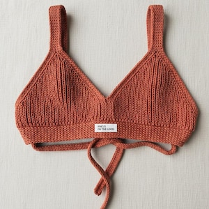 May include: A rust-colored knitted top with a V-neck design and tie straps. The top features a small rectangular tag with the text "FOCUS ON THE GOOD". The straps are wide and the knit has a ribbed texture.