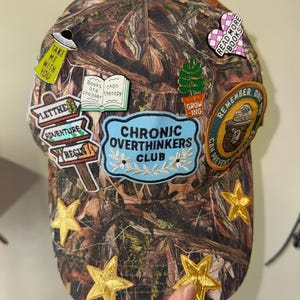 Könnte beinhalten: Tarnfarbene Baseballkappe mit Aufnähern und Anstecknadeln. Aufnäher mit Text wie "Chronic Overthinkers Club", "Let the Adventure Begin" und "Read More Books". Goldene Sternverzierungen sind ebenfalls vorhanden.