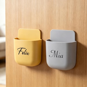 Peut inclure: Deux poches de rangement murales, une jaune et une gris clair, chacune dotée d'un panneau arrière plat et d'un compartiment ouvert arrondi. La poche jaune porte le nom Felix en écriture noire, et la poche grise porte le nom Mia en écriture noire.