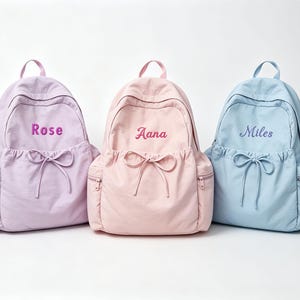 Pode incluir: Três mochilas nas cores lavanda, rosa claro e azul claro. Cada bolsa possui um bolso frontal com cordão, uma alça superior e nomes bordados: Rose na bolsa lavanda, Aana na bolsa rosa e Miles na bolsa azul.