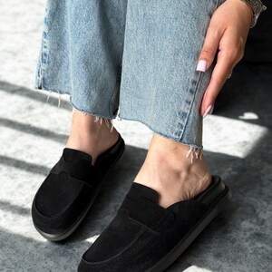 May include: Black suede slip-on loafers with a rounded toe and a low, dark gray sole. The shoes have a decorative strap across the top and are worn with light blue denim jeans.