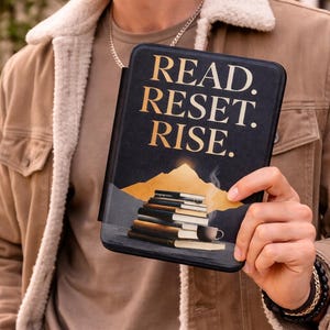 Könnte beinhalten: Schwarze E-Reader-Hülle mit goldfarbenem Schriftzug „READ. RESET. RISE.“ Die Hülle zeigt eine Berglandschaft mit einem Stapel Bücher und einer Kaffeetasse. Die Hülle wird von einer Person gehalten, die eine braune Jacke trägt.