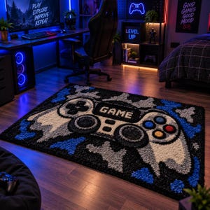 May include: A rectangular area rug featuring a tufted design of a video game controller in shades of black, white, gray, and blue. The center of the controller displays the word GAME in white letters, surrounded by blue and gray camouflage patterns.