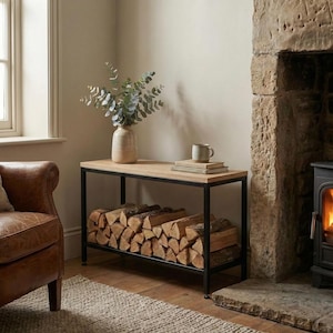 Op de afbeelding: Een houten en zwart metalen houtopslagrek. De bovenste plank bevat een vaas met groen, een mok en boeken. De onderste plank is gevuld met brandhout. De kamer heeft een open haard en een leren fauteuil.