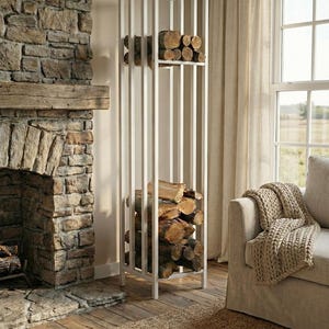 Puede incluir: Un leñero blanco vertical con dos estantes, lleno de leña. El leñero está junto a una chimenea de piedra y un sofá beige con una manta de punto. La habitación tiene una paleta de colores neutros. El leñero mide unos 100 cm de ancho.