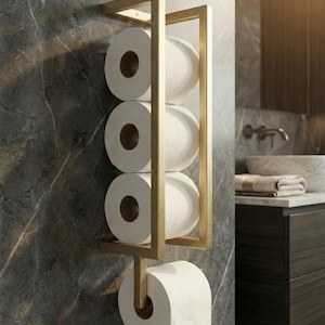 May include: A gold-colored metal toilet paper holder mounted on a gray marble wall. The holder has a rectangular frame with four rolls of white toilet paper and one roll dispensing from the bottom. The bathroom fixture adds a touch of elegance.