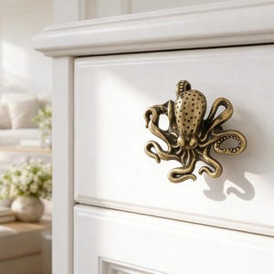 May include: A bronze octopus-shaped drawer pull is mounted on a white cabinet. The detailed pull features a textured body and curling tentacles. The cabinet is part of a larger white furniture set, with a glimpse of a living room in the background.