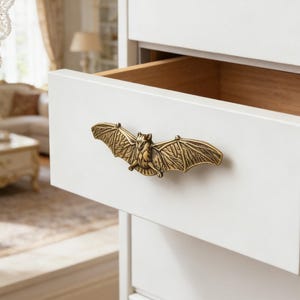 May include: A white chest of drawers with an open drawer. The drawer features a brass bat-shaped handle. The handle is intricately detailed. The interior of the drawer is made of wood. The room is bright and airy.