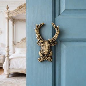 May include: A brass door knocker in the shape of a deer head with large antlers, mounted on a light blue door. The knocker has intricate details and a decorative collar. The background shows a glimpse of a bedroom with a white bed.