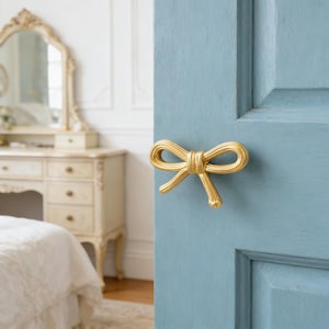 May include: A gold-colored bow-shaped door handle is mounted on a light blue door. The handle has a textured surface and is in a bedroom setting with a vanity and bed in the background.
