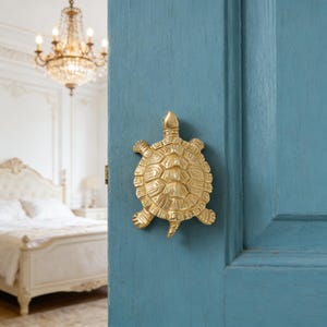 Puede incluir: Un llamador de puerta dorado en forma de tortuga montado en una puerta azul. El llamador tiene un caparazón y aletas texturizadas. El fondo muestra un dormitorio con una lámpara de araña y una cama blanca.