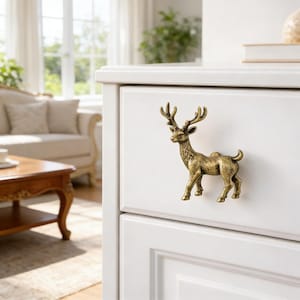 May include: A brass-colored deer-shaped cabinet knob is mounted on a white drawer. The deer has detailed antlers and a standing pose. The knob adds a decorative touch to furniture.
