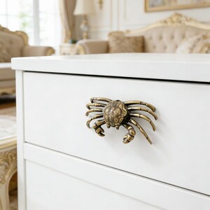 Op de afbeelding: Een witte lade met een bronzen krabvormige handgreep. De handgreep is gedetailleerd met scharen, poten en een getextureerd lichaam. De lade maakt deel uit van een witte kast, met een glimp van een kamer op de achtergrond.